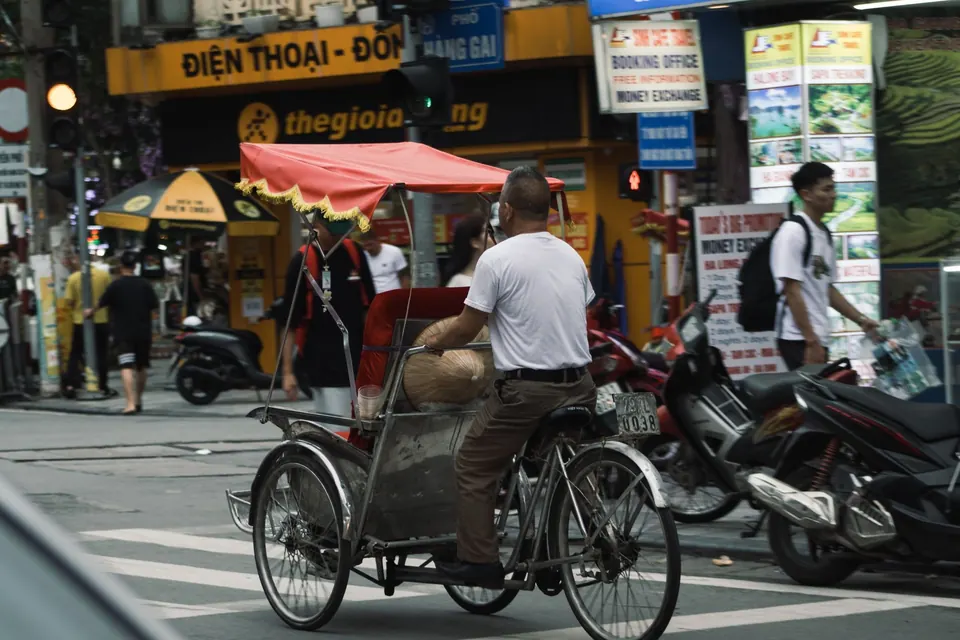 A red cyclo pedicab crossing a busy street in the Old Quarter of Hanoi among scooters and shop signs