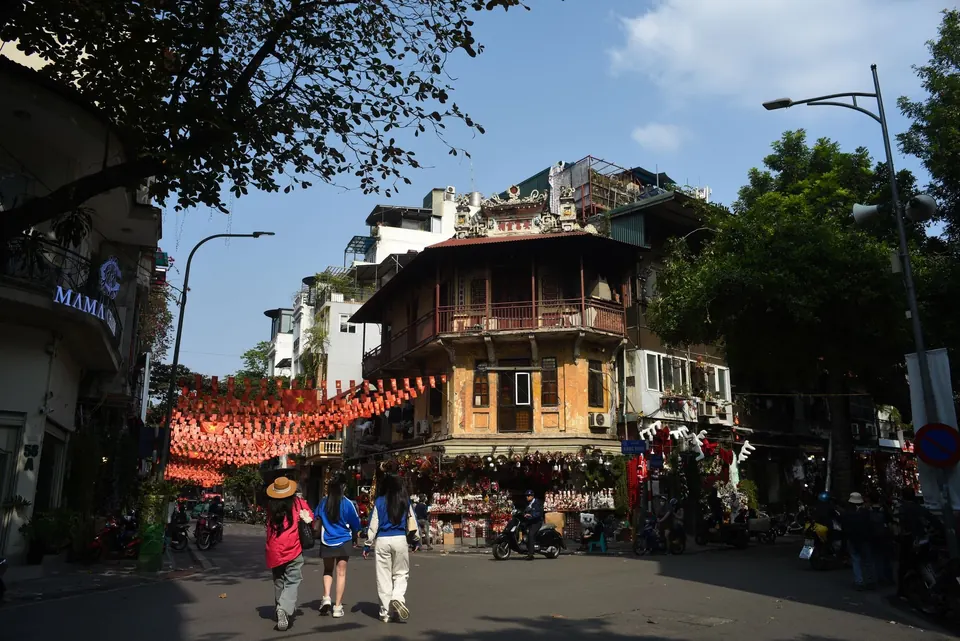 Strings of red lanterns hung across an Old Quarter street corner in Hanoi, with an old tiled corner house and people walking below