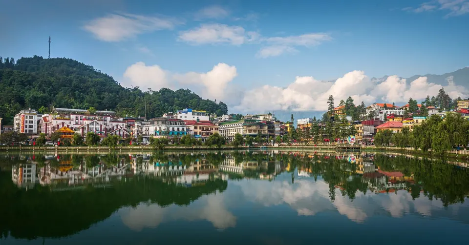 Sapa town reflected in Sapa lake on a clear morning, with hotel rooftops along the shore and a forested hill behind